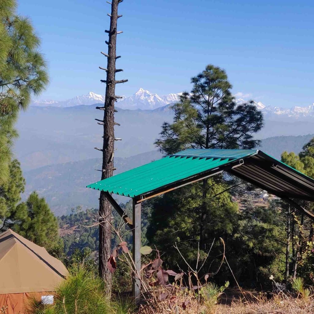 A luxury swiss tent with a peach colored roof and a wooden porch at Him 180, a resort in Ranikhet that offers adventure glamping with breathtaking views of the Himalayas. The tent is surrounded by lush greenery and has a clear view of the snow-capped mountains in the distance. The photo was taken during the day and the lighting is natural.