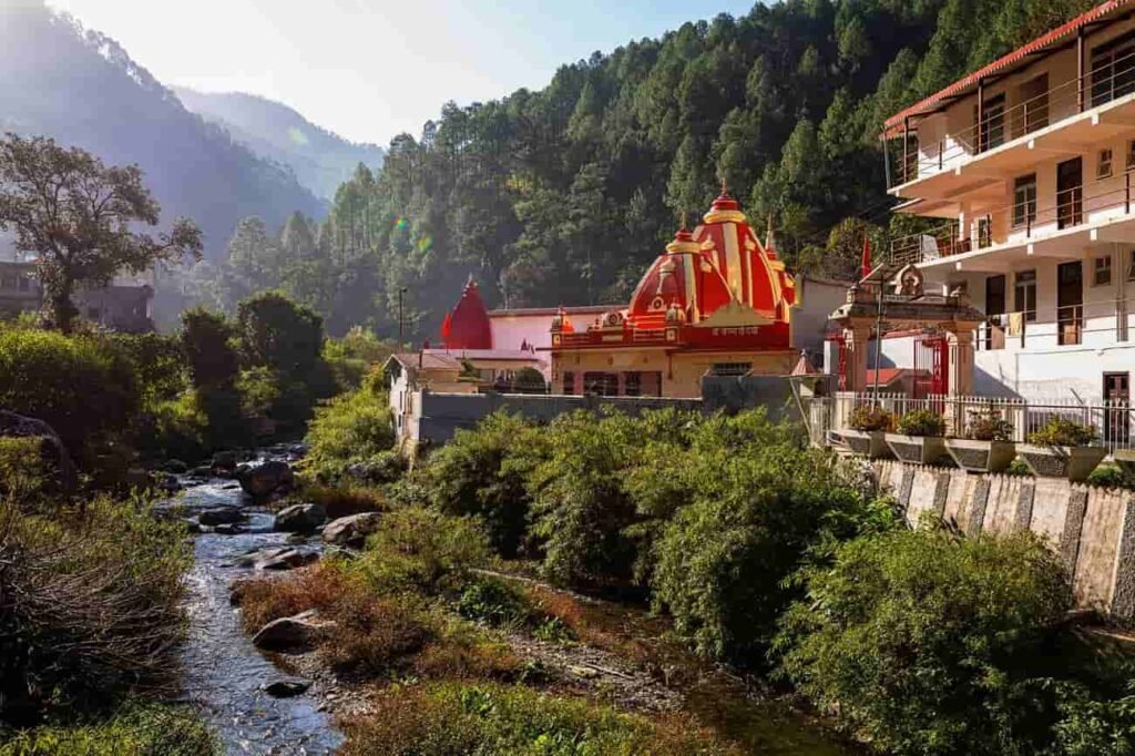 A view of Kainchi Dham Ashram and Hanuman Temple surrounded by green hills and trees with a river flowing beside it.