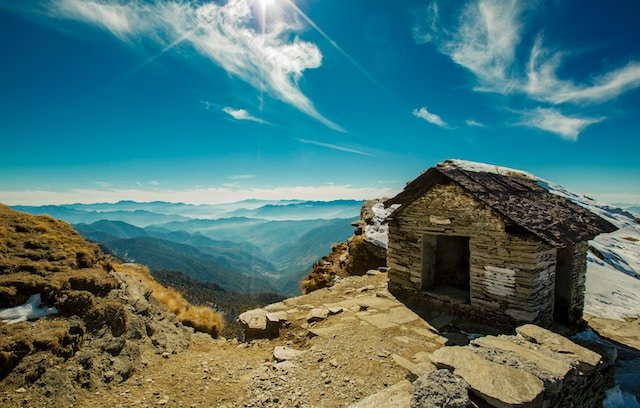 A stunning view of Chandrashila, a summit in Uttarakhand, India, where Lord Rama meditated after killing Ravana. The image shows a stone hut on the top of the mountain, surrounded by snow and rocks, with a clear blue sky and the Himalayan peaks in the background. This image depicts one of the off-the-beaten-path travel spots in Uttarakhand that most tourists don’t know about.
