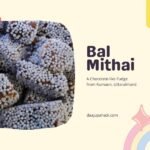 A close up of bal mithai, a traditional Indian sweet from Kumaon, Uttarakhand, that looks like chocolate fudge covered in white sugar balls. The image has a pale yellow background with pink and blue illustrations of a cupcake and text that says “Bal Mithai, A Chocolate-like Fudge from Kumaon, Uttarakhand” and “dajuaupahadi.com”.
