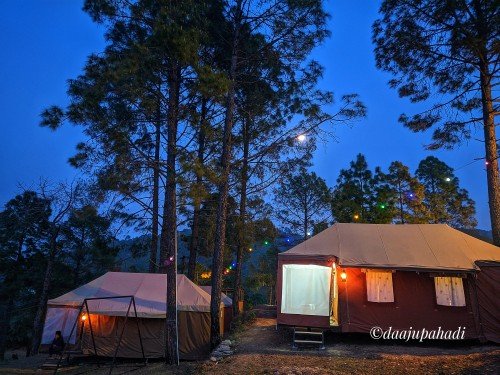 Him 180 campsite in the woods at night with two swiss cottage tents and fairy lights. Tents have lights on inside. The tents are on a clearing surrounded by tall trees. The moon and the blue sky are visible through the trees. The photo is credited to @aajaypahadi.